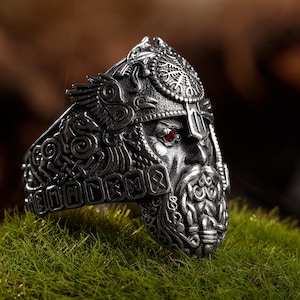 May include: A detailed silver ring featuring a Viking warrior with a helmet, beard, and red gemstone eyes. The ring has intricate carvings and is set against a blurred green and brown background.