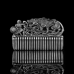 May include: A silver-colored comb with a detailed Viking design. The comb features a warrior with a shield and sword facing a snarling wolf. Intricate knotwork and vertical teeth complete the design, reflecting in the dark background.