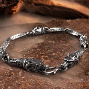 May include: A silver bracelet with two wolf head charms. The bracelet is made of a chain link design with a detailed pattern.