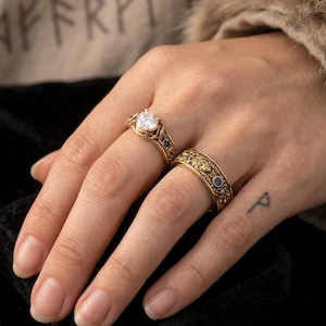 May include: Two gold rings with intricate designs. One ring features a large, clear gemstone, while the other has a patterned band with small blue stones. The rings are displayed on a hand.