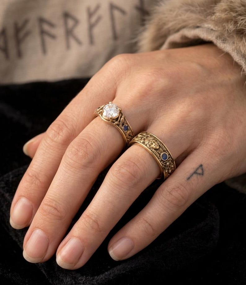 May include: Two gold rings with intricate designs. One ring features a large, clear gemstone, while the other has a patterned band with small blue stones. The rings are displayed on a hand.