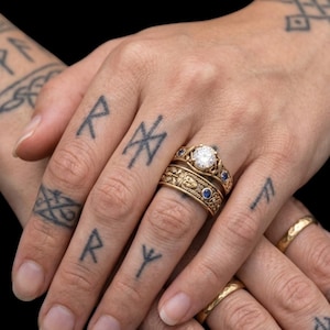 May include: Close-up of multiple gold rings, including a diamond engagement ring and a band with blue gemstones. The rings are on a hand with tattoos of runes and other symbols. The background is black.