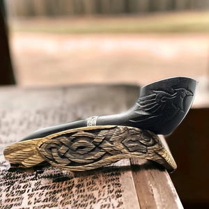 Op de afbeelding: Een zwarte, gesneden tabakspijp met een vogelontwerp op de kop, rustend op een houten standaard met Keltische knoopversieringen. De pijp heeft een zilveren band bij het mondstuk. De standaard is gemaakt van lichtgekleurd hout.