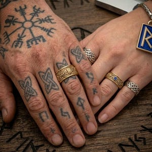 May include: Close-up of hands adorned with gold and silver rings, featuring intricate Celtic knot designs and a blue gemstone. The hands are tattooed with Viking symbols, resting on a wooden surface with carved runes. A pendant with a blue enamel inlay is also visible.