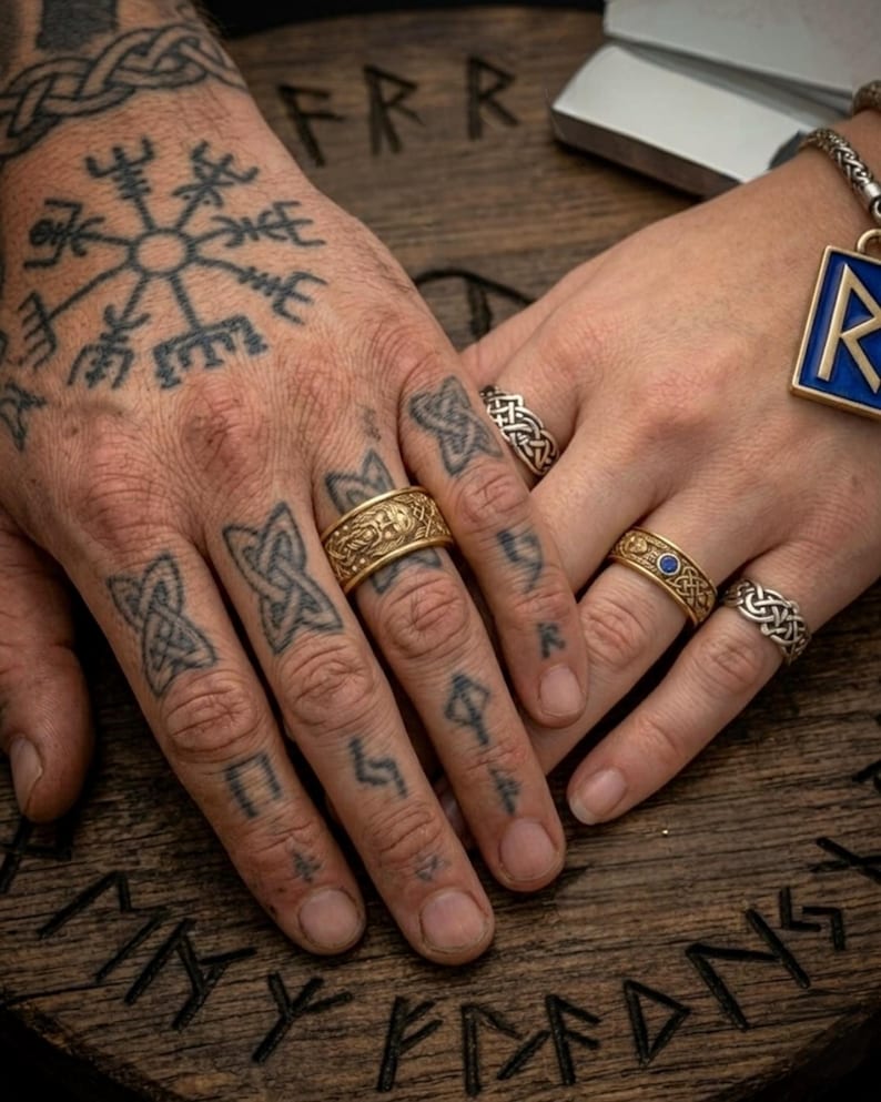 May include: Close-up of hands adorned with gold and silver rings, featuring intricate Celtic knot designs and a blue gemstone. The hands are tattooed with Viking symbols, resting on a wooden surface with carved runes. A pendant with a blue enamel inlay is also visible.