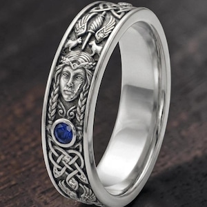 May include: Silver-toned ring with intricate Celtic knot and face detailing. Features a blue gemstone and detailed carvings of figures and patterns. The ring's design is inspired by Celtic and Viking themes.