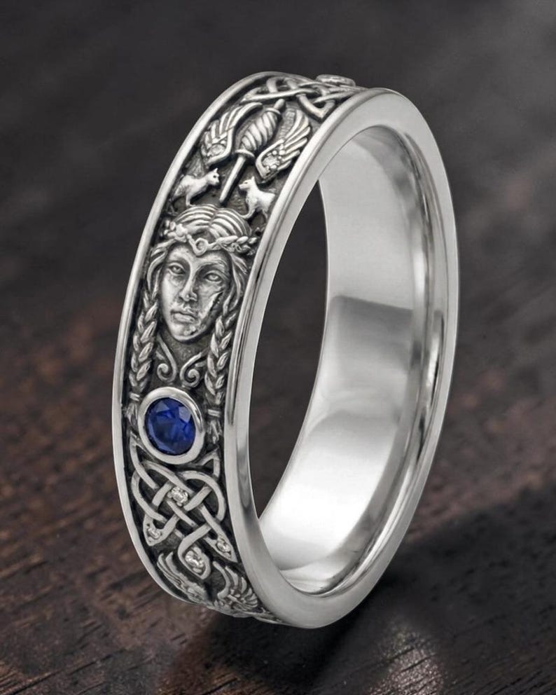 May include: Silver-toned ring with intricate Celtic knot and face detailing. Features a blue gemstone and detailed carvings of figures and patterns. The ring's design is inspired by Celtic and Viking themes.