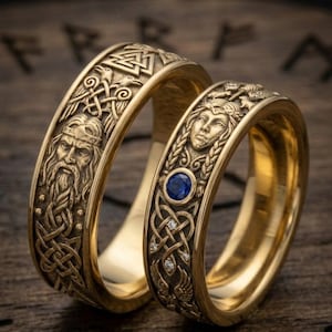 May include: Two gold-colored rings with intricate Viking-inspired designs. One ring features a bearded face and knotwork, the other has a blue gemstone and a female face. The rings are set against a wooden background.