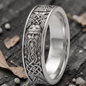 May include: A silver-colored ring with a detailed Viking design. The ring features a carved image of a bearded man, intricate knotwork, and other Viking symbols. The ring has a polished finish and a wide band.