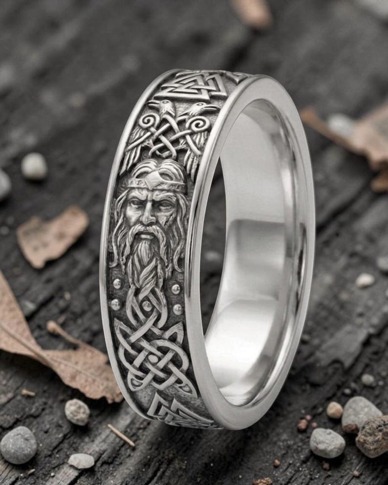 May include: A silver-colored ring with a detailed Viking design. The ring features a carved image of a bearded man, intricate knotwork, and other Viking symbols. The ring has a polished finish and a wide band.