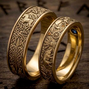 May include: Two gold-colored rings with intricate embossed designs. One ring features a horse and knotwork, the other a boar, wheat, and a wheel. The rings have a polished finish and are set against a dark wooden surface.