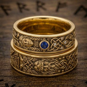 May include: Two gold-colored rings stacked on a wooden surface. The rings feature intricate Celtic knotwork and engraved figures. One ring has a blue gemstone and small diamonds. The rings are a symbol of Viking culture.