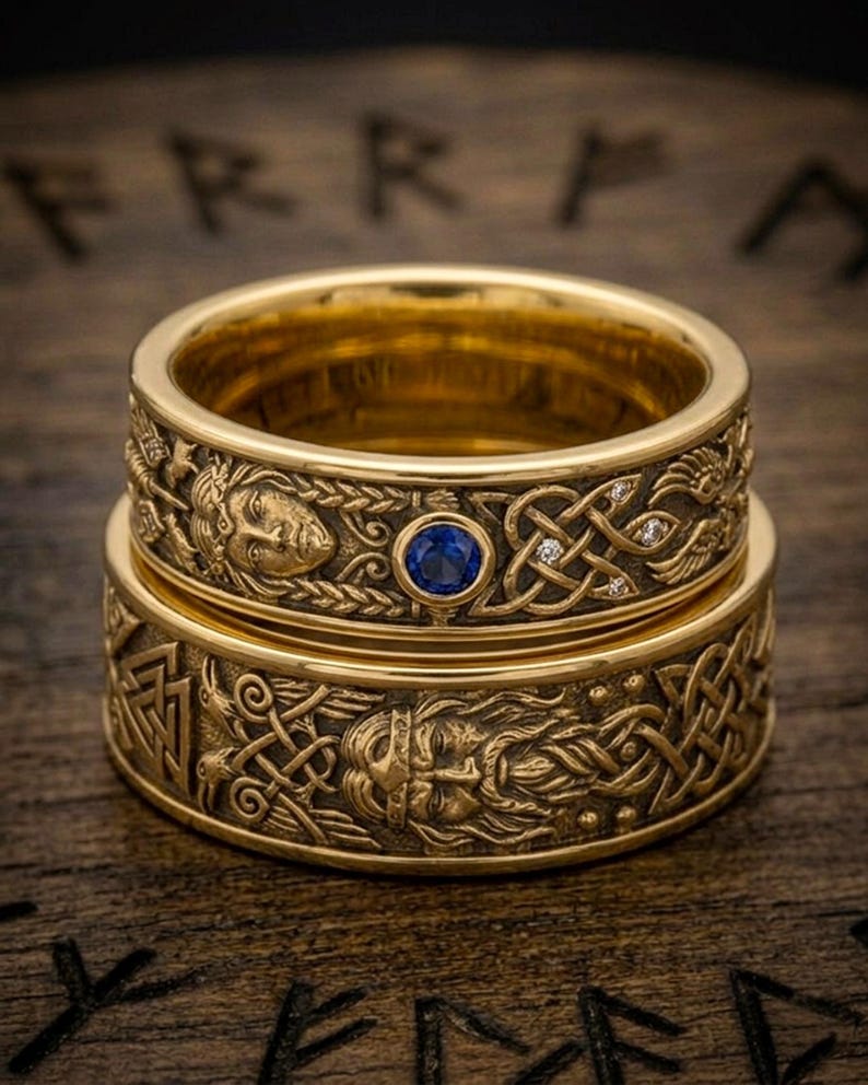 May include: Two gold-colored rings stacked on a wooden surface. The rings feature intricate Celtic knotwork and engraved figures. One ring has a blue gemstone and small diamonds. The rings are a symbol of Viking culture.