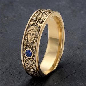 May include: A gold-colored ring with intricate Celtic knot and figure detailing. The ring features a central blue gemstone and small accent diamonds. The design includes a woman's face and other symbolic elements.