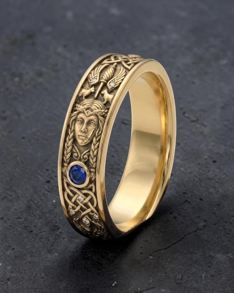 May include: A gold-colored ring with intricate Celtic knot and figure detailing. The ring features a central blue gemstone and small accent diamonds. The design includes a woman's face and other symbolic elements.