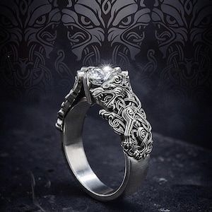 May include: A silver ring with a detailed wolf design. The ring holds a sparkling, clear gemstone, secured by intricate metalwork. The band and setting are decorated with Celtic knot patterns, creating a unique and eye-catching piece of jewellery.