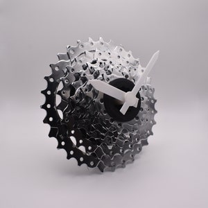 Könnte beinhalten: Eine silberne Fahrradkassettenuhr mit weißen Zeigern. Das Zifferblatt der Uhr ist schwarz.