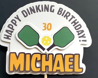 May include: White cake topper with the text "Happy Dink'n Birthday!" and "Michael" in orange. The topper features two green pickleball paddles with a yellow pickleball in the centre. The number 30 is written in orange on the topper.