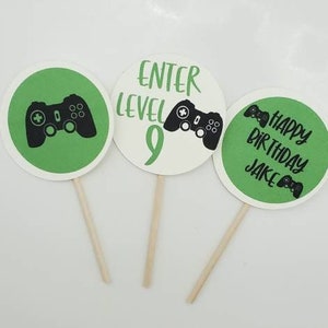 May include: Three green and white round cupcake toppers with black game controller graphics. The toppers say "Enter Level 9", "Happy Birthday Jake" and a game controller graphic.