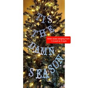 May include: A silver glitter Christmas banner that reads "Tis the Damn Season" hanging on a Christmas tree with white lights. The letters range in size from 1.5 to 5 inches.