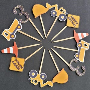 May include: A collection of birthday cake toppers featuring construction-themed designs. The toppers include yellow construction vehicles, orange traffic cones, the number 3, and signs that read "Happy Birthday". The toppers are arranged on wooden sticks.
