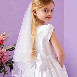May include: A girl in a white satin dress and a veil. The dress has a bow at the back and a simple design. The veil has a beaded trim. The background is a soft purple. The image is well-lit and the girl is looking at the camera.
