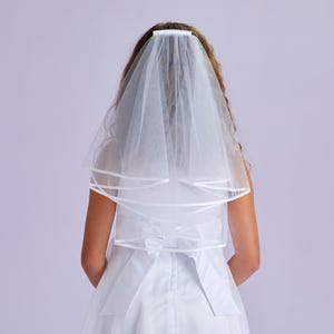 May include: A white veil with a layered design and satin ribbon trim. The veil is attached to a white headband and worn over a white dress. The back of the dress features a bow.