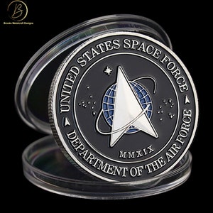 May include: A silver-colored coin with a black background featuring the United States Space Force logo. The logo includes a delta symbol, a globe, and the text "United States Space Force" and "Department of the Air Force". The year "MMXIX" is also included on the coin.
