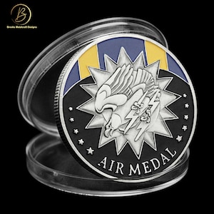 May include: A silver coin with a black background and a silver eagle with wings spread. The eagle is holding a lightning bolt in its talons. The coin has a blue and yellow ribbon around the edge and the words "AIR MEDAL" inscribed on the bottom.