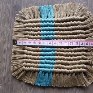 May include: A woven square coaster with a blue stripe down the centre. The coaster is made of natural fibres and has a fringe border. The coaster measures 15 cm wide.