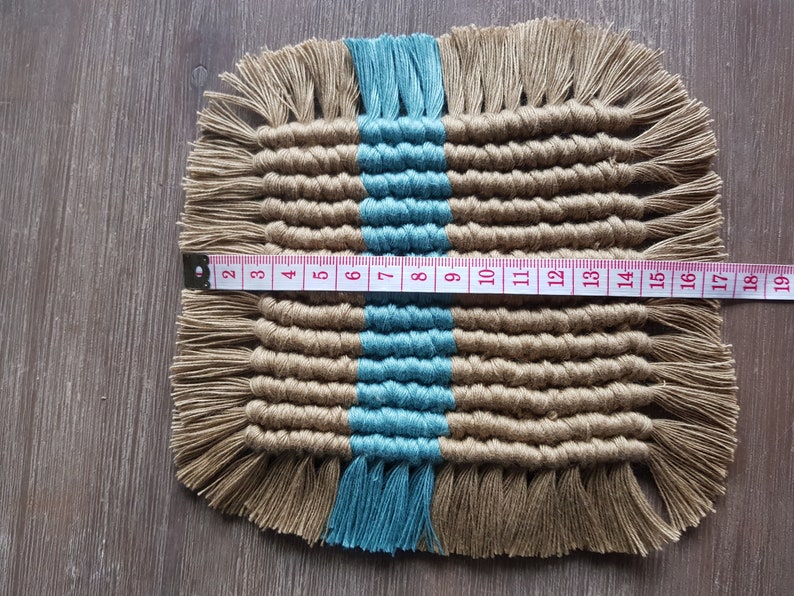 May include: A woven square coaster with a blue stripe down the centre. The coaster is made of natural fibres and has a fringe border. The coaster measures 15 cm wide.