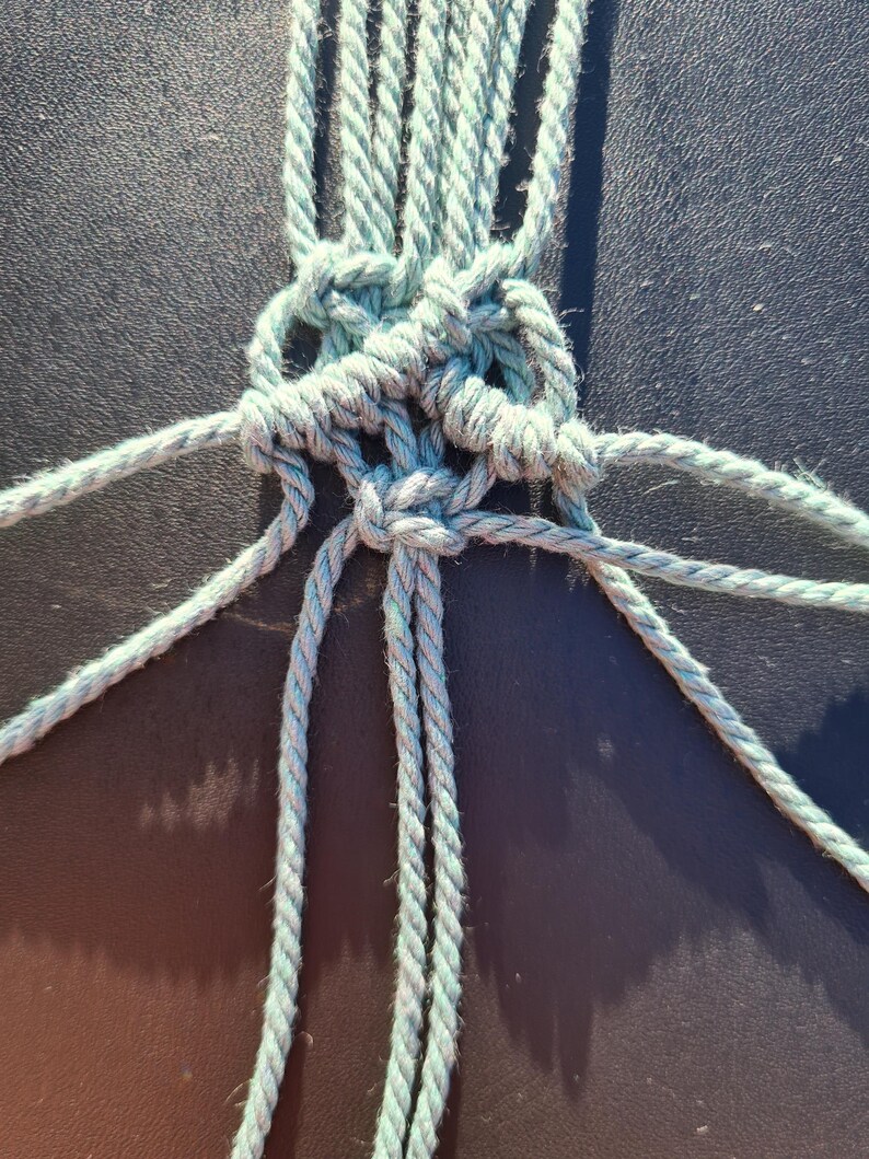 K&ouml;nnte beinhalten: Nahaufnahme eines Makramee-Knotens in Hellt&uuml;rkis. Der Knoten ist aufwendig mit mehreren Seilstr&auml;ngen gewebt und erzeugt ein strukturiertes Muster. Der Hintergrund ist dunkel und einfarbig und bildet einen Kontrast zum Knoten.