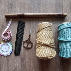 May include: A set of craft supplies for macrame projects, including two spools of rope, one in beige and one in teal, a wooden dowel, a measuring tape, a comb, a pincushion with colourful pins, and a pair of scissors.