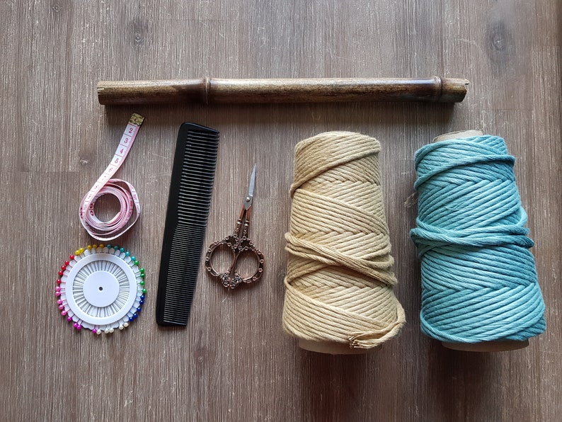 May include: A set of craft supplies for macrame projects, including two spools of rope, one in beige and one in teal, a wooden dowel, a measuring tape, a comb, a pincushion with colourful pins, and a pair of scissors.