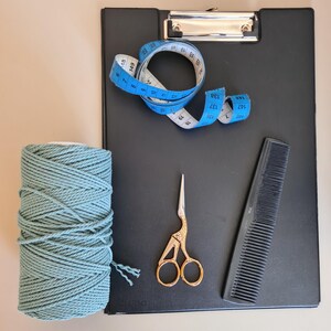 K&ouml;nnte beinhalten: Ein schwarzes Klemmbrett mit einem blauen Ma&szlig;band, einer Spule aus teal-farbenem Makramee-Garn, einer goldenen Schere und einem schwarzen Kamm. Das Ma&szlig;band ist in Zentimetern markiert.