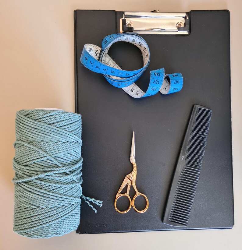 K&ouml;nnte beinhalten: Ein schwarzes Klemmbrett mit einem blauen Ma&szlig;band, einer Spule aus teal-farbenem Makramee-Garn, einer goldenen Schere und einem schwarzen Kamm. Das Ma&szlig;band ist in Zentimetern markiert.