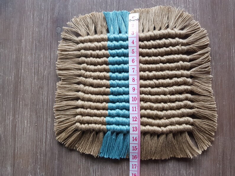 May include: A square woven coaster with a blue and brown colour scheme. The coaster is approximately 15 cm wide and has a fringe border.