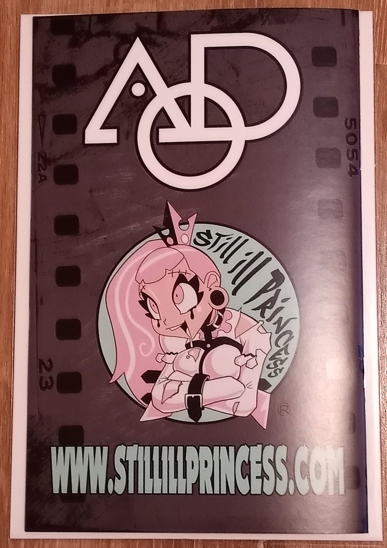 May include: A black and white illustration of a stylized letter "A" and a circle with a small dot inside. Below the illustration is a pink cartoon character with a crown, wearing a white shirt and black straps. The character has pink hair and is holding their arms crossed. The text "Still Ill Princess" is written in a circle around the character. The text "www.stillillprincess.com" is written below the character.