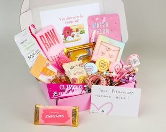 Pode incluir: Uma caixa de presente rosa cheia de vários itens, incluindo um esfoliante para os pés, uma máscara facial, uma barra de chocolate, um colar e um bilhete escrito à mão que diz "Sua mensagem personalizada aqui!" A caixa é decorada com bolinhas rosa e brancas e as palavras "Você merece ser mimado!"