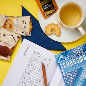 K&ouml;nnte beinhalten: Eine Draufsicht auf eine gelbe Oberfl&auml;che mit einer Tasse Tee, einem Teller mit Butterkeksen, zwei Packungen Kekse, einem Bleistift, einem Sudoku-Puzzlebuch und einem Kreuzwortr&auml;tselbuch. Das Kreuzwortr&auml;tselbuch tr&auml;gt den Text "Best Ever Crosswords Unleash your inner genius".