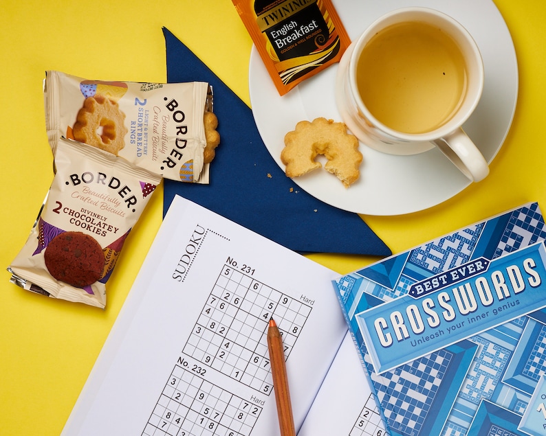 K&ouml;nnte beinhalten: Eine Draufsicht auf eine gelbe Oberfl&auml;che mit einer Tasse Tee, einem Teller mit Butterkeksen, zwei Packungen Kekse, einem Bleistift, einem Sudoku-Puzzlebuch und einem Kreuzwortr&auml;tselbuch. Das Kreuzwortr&auml;tselbuch tr&auml;gt den Text "Best Ever Crosswords Unleash your inner genius".