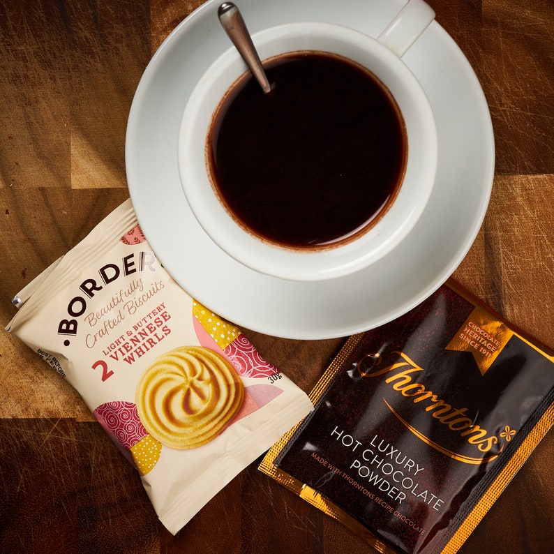 K&ouml;nnte beinhalten: Eine Tasse hei&szlig;e Schokolade mit einem L&ouml;ffel darin, eine Packung mit leichten und butterartigen Wiener Wirbeln und eine Packung Thorntons Luxury Hot Chocolate Powder. Die Packung mit dem Schokoladenpulver ist goldfarben mit schwarzem Text und dem Thorntons-Logo.