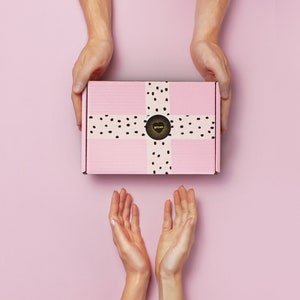 K&ouml;nnte beinhalten: Zwei H&auml;nde reichen nach einem rosafarbenen Geschenkbox mit einem wei&szlig;en Band und schwarzen Punkten. Die Box hat einen goldenen Herzaufkleber mit dem Wort "Love" darauf.