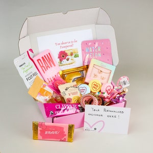 May include: Pink and white gift box filled with various self-care items, including face masks, foot peel, bath bombs, and a chocolate bar. The box also includes a handwritten note that says "Your Personalised Message Here!"