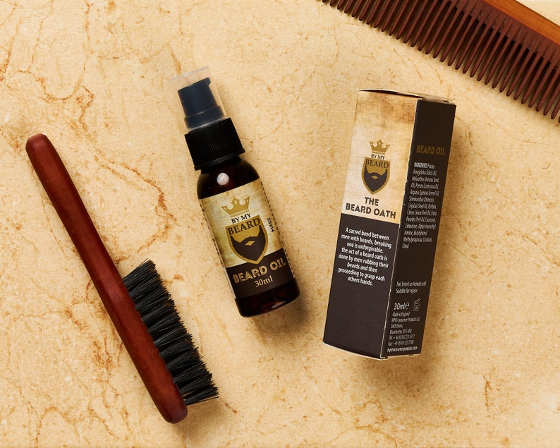 May include: A brown wooden comb, a brown wooden brush, and a glass bottle of beard oil with a gold label that says "By My Beard The Beard Oath" and "Beard Oil 30ml".