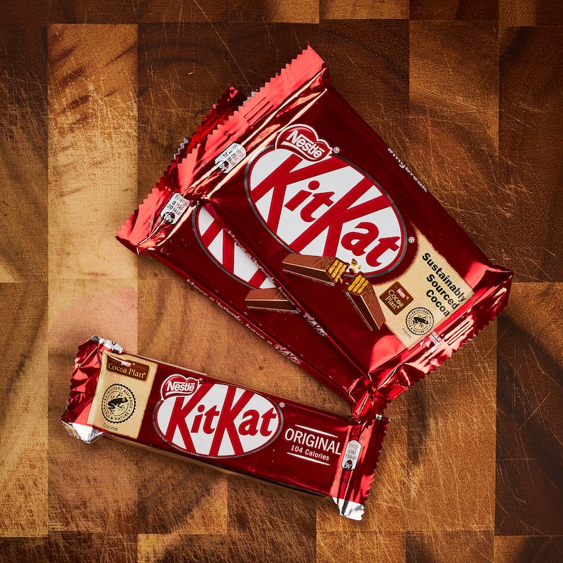 K&ouml;nnte beinhalten: Zwei rote und wei&szlig;e Kit Kat Schokoladenriegelverpackungen mit dem Text "Nestle Kit Kat" und "Nachhaltig Bezogener Kakao". Eine kleinere rote und wei&szlig;e Kit Kat Verpackung mit dem Text "Original 164 Kalorien" befindet sich im Vordergrund.
