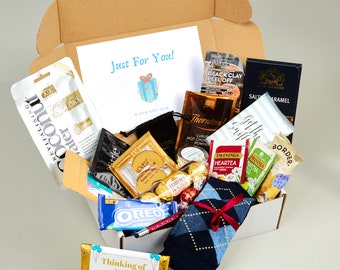 May include: A brown cardboard box filled with various gifts, including a pair of blue and white socks, a bag of chocolate, a box of tea, a box of face masks, and a card that says "Just For You!" and "Thinking of You!"