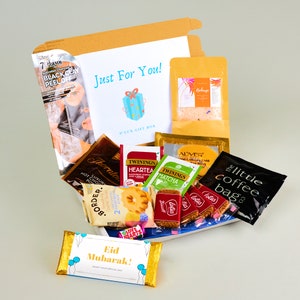 May include: A gift box filled with various items, including tea bags, coffee, cookies, and a chocolate bar. The box is decorated with a blue and white gift box design and the text "Just For You!" The box also includes a small card with the text "Eid Mubarak!" and a gold ribbon.