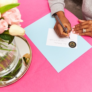 May include: A person is writing on a white card with a blue background. The card has a gold sticker with a black design on it. The text on the card reads "You're remarkable. I know you feel happy".