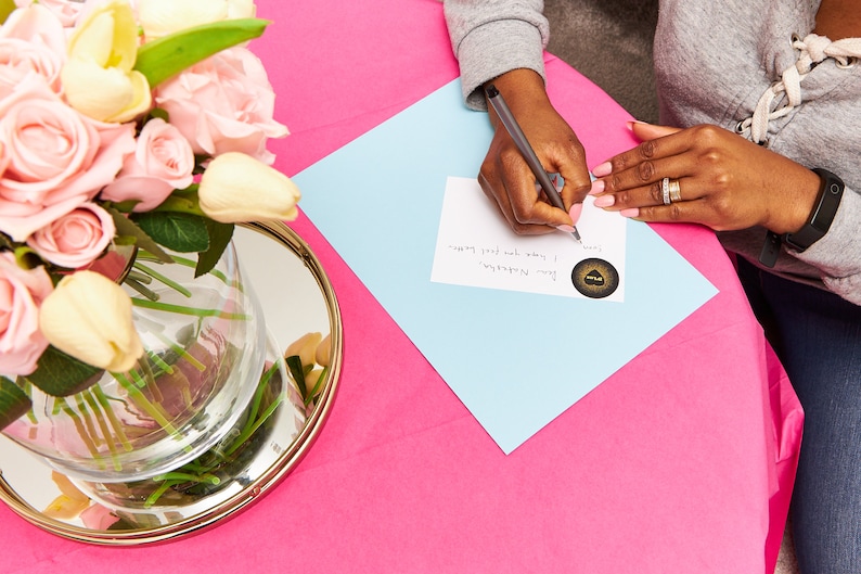 May include: A person is writing on a white card with a blue background. The card has a gold sticker with a black design on it. The text on the card reads "You're remarkable. I know you feel happy".